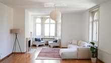 Bright living room with white walls, wooden flooring, large windows, a fireplace, a white sectional sofa, two white armchairs, a small wooden table, a floor lamp, a pendant lamp, and a potted plant.