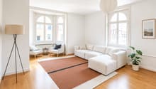 Bright living room with large arched windows, white walls, light wood flooring, a sectional sofa with an ottoman, two armchairs, a small side table, a floor lamp, a potted plant, and a picture frame on the wall.