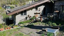 A rustic garden area with a small house, potted plants, outdoor furniture, and a stone pathway.