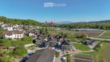 Aerial view of a rural village with traditional houses, a church, and a construction site with a crane in the background, surrounded by green fields and mountains.