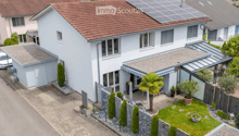 House. A two-story white house with a red-tiled roof, solar panels on the roof, and a modern garage. The house features large windows and a well-maintained garden with a palm tree, shrubs, and a small lawn.