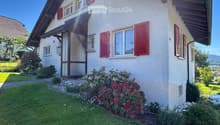 Two-story house with white walls, red shutters, and a well-maintained garden with flowering shrubs and green grass.