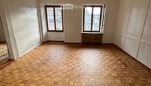 Empty room with wooden herringbone flooring, white walls, and two windows with brown frames. The ceiling has exposed wooden beams.