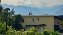 A two-story yellow house with white-framed windows and a gray roof, surrounded by lush greenery and palm trees, with a scenic mountain backdrop.