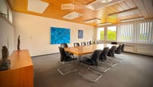 Conference room with a long wooden table surrounded by black chairs, a sculpture on a side table, and artwork on the walls. Large windows provide natural light.