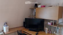 A small, furnished room with a wooden table, a black chair, a large screen, a remote control, a small cabinet, and various items on the shelves and table.