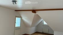 Empty attic room with wooden flooring, white walls, slanted ceiling, and two windows.