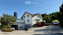 Two-story house with white exterior, multiple windows, garage, and balcony. There are two cars parked in the driveway and a silo in the background.