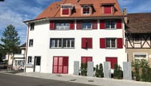 House. Two-story house with white walls, red shutters, and a red-tiled roof. Features multiple windows, a front door, and small stone planters with greenery.