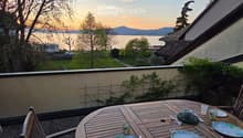 Balcony with a wooden table set for two, overlooking a lake and mountains at sunset.