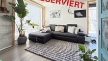 Modern living room with a grey sectional sofa, large shaggy rug, potted plants, and artwork on the walls.