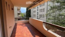 Wooden deck balcony with glass railing, beige awning, and view of neighboring buildings and greenery.