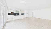 Bright, open-plan living and kitchen area with white tiled flooring, white kitchen cabinets, a central island, and modern appliances.