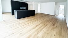 Empty room with light wooden flooring and white walls, featuring a dark kitchen island with cabinets and a sink.