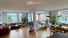 Open-plan living and dining area with wooden flooring, large windows, and a balcony. Features a red sofa, dining table with chairs, and a kitchen counter with a faucet and various items.