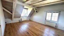 Empty attic room with wooden floors, white walls, exposed wooden beams, and two windows with blinds.