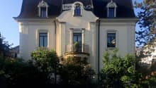 A three-story white house with ornate architectural details, including decorative window surrounds and a small balcony with a potted plant. The house features multiple windows and is surrounded by lush greenery and flowering bushes.