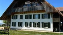 House. Traditional two-story house with white walls, dark green shutters, and a wooden balcony on the upper floor. The roof is red-tiled with a prominent wooden overhang. The surrounding area includes a lawn, a small seating area with stone blocks and a m