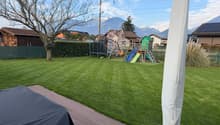 Well-maintained lawn with a wooden deck, covered grill, and a children's play area including a slide and swing set. The background features a hedge, trees, and distant mountains.