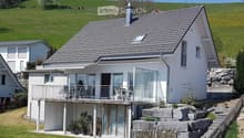 A modern two-story house with a grey tiled roof, white exterior, and a spacious balcony with glass railings. The house is surrounded by a well-maintained garden with potted plants and a stone wall.