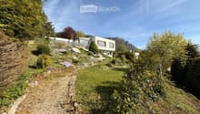 A modern house with a raised ground floor, surrounded by a landscaped garden with a gravel path, lush greenery, and a scenic mountain view in the background.