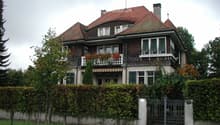 Two-story house with red-tiled roof, multiple windows, balconies, and surrounding greenery.