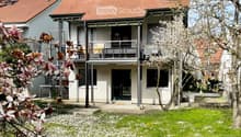 Two-story modern house with red-tiled roof, large windows, and a balcony. The front yard features a well-maintained lawn with white flowers and blooming trees.