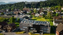 Aerial view of a modern building with solar panels on the roof, surrounded by a small garden and integrated into a village setting with traditional houses and green spaces.