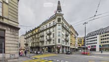 A multi-story building with a distinctive tower and spire, located at a street corner with a pedestrian crossing.