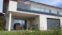 Modern two-story house with a large glass balcony on the upper level, a covered patio area with a grill, and a well-maintained garden.
