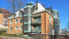 A multi-story brick building with green balconies and a snow-covered roof, situated on a hillside with a curved road in front.