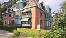 A multi-story brick house with green balconies, multiple windows, and a well-maintained hedge in front.