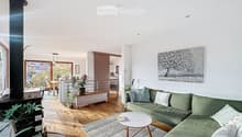Modern living room with a green sectional sofa, white coffee table, fireplace, artwork, and large windows.