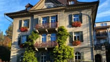 Three-story residential house with beige facade, multiple windows, balconies, and decorative railings. Greenery and flower boxes adorn the building.