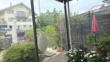 A well-maintained garden area with wooden decking, potted plants, a small tree, and a pink umbrella. The space is enclosed by glass walls, with a view of neighboring houses and greenery.