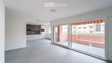 Modern, unfurnished living space with a fully equipped kitchen area, large sliding glass doors leading to a balcony, and grey tiled flooring.
