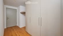 A narrow hallway with light wooden flooring, white walls, built-in white cabinets with silver handles, and a closed door at the end.