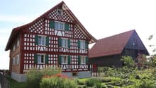 Traditional half-timbered house with red and white exterior, green shutters, and a well-maintained garden with various plants and shrubs.