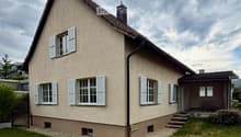 Two-story house with beige siding, white shutters, and a small porch with a table and chairs.
