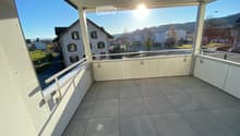 Balcony with tiled floor, white railings, and a view of a residential area with houses and greenery.
