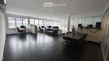 Modern office space with black flooring, large windows, office desks, chairs, a sofa, and plants.