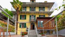 A two-story yellow building with green shutters and balconies, featuring a staircase leading to the entrance and a small wooden annex nearby.