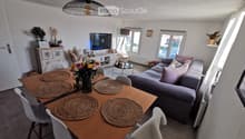 Modern living and dining area with a grey sofa, wooden dining table with six chairs, a television, and decorative items including a flower vase and a floor lamp.