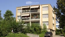Four-story residential building with balconies, awnings, and flower boxes, surrounded by greenery and a parking area.