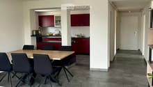 Open-plan living and dining area with grey tiled flooring, a wooden dining table surrounded by black chairs, and a modern kitchen with red cabinets and stainless steel appliances.