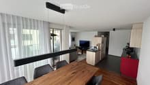 Open-plan living and dining area with a wooden dining table, chairs, a television, a small kitchen area with a sink, refrigerator, and a balcony with sheer curtains.