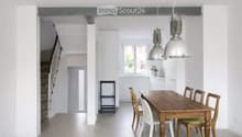 Bright, open-plan dining area with a wooden dining table, white and wooden chairs, pendant lights, and a small stool near the kitchen area.
