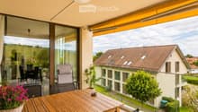 Balcony with wooden table, green chairs, and a flower pot, overlooking a residential area with houses and greenery.
