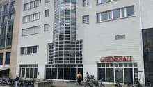 Modern multi-story building with white facade, large windows, and a cylindrical tower section. The ground floor has glass storefronts with signage reading "GENERALI" and "WEIGTIKLASSE 11". Bicycles are parked along the sidewalk in front of the building.