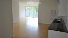Empty room with wooden flooring, white walls, and a small kitchen area with a sink and cabinets. Large sliding glass doors lead to a balcony.